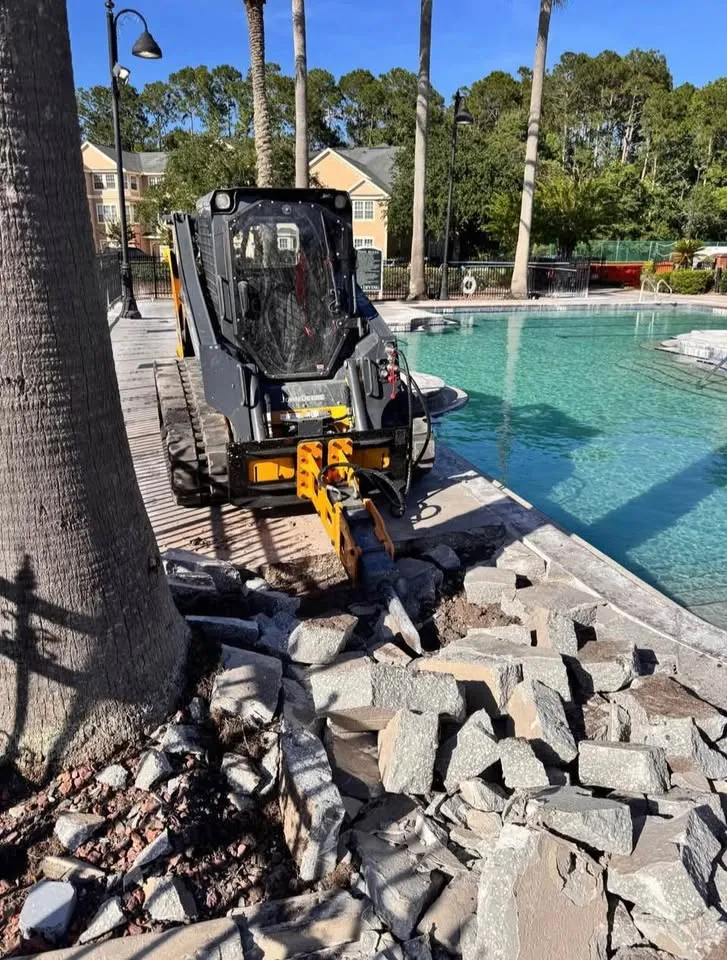 Track loader with a breaker working along a pool edge, broken slabs stacked in the foreground