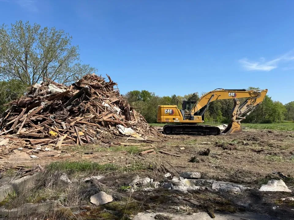Excavator beside a stacked pile of timber debris on a flattened residential lot