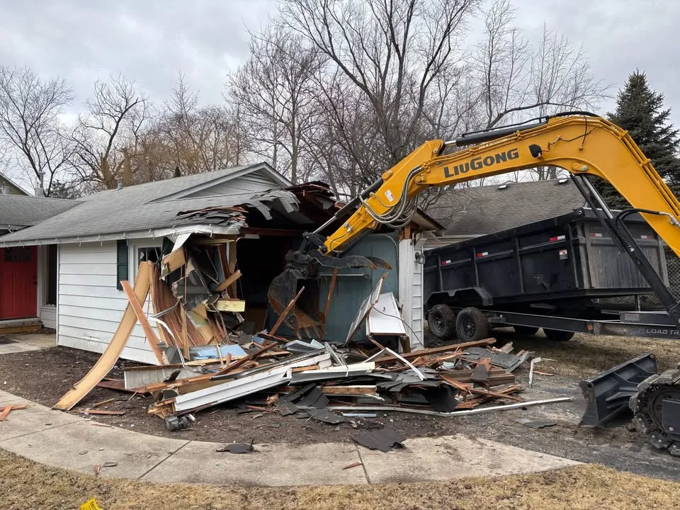 Excavator arm tearing the roof off a single-storey bungalow next to a dump trailer