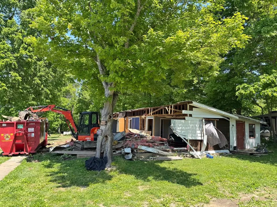 Excavator and red roll-off bin in front of a partly demolished bungalow under a large shade tree