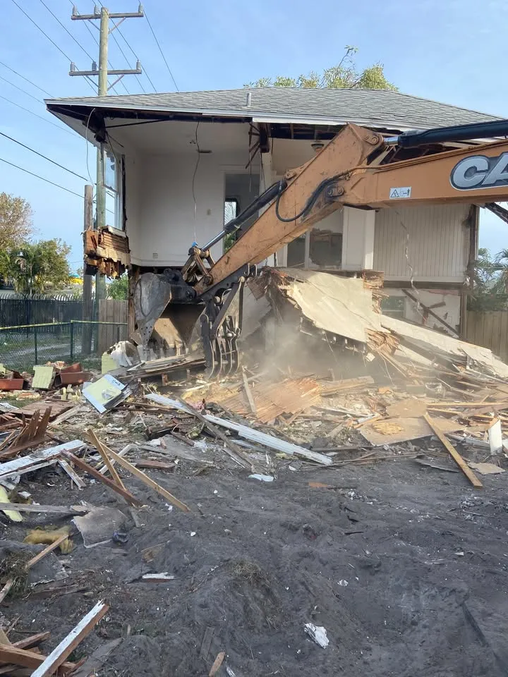 Excavator bucket breaking through the front wall of a two-storey house with dust rising