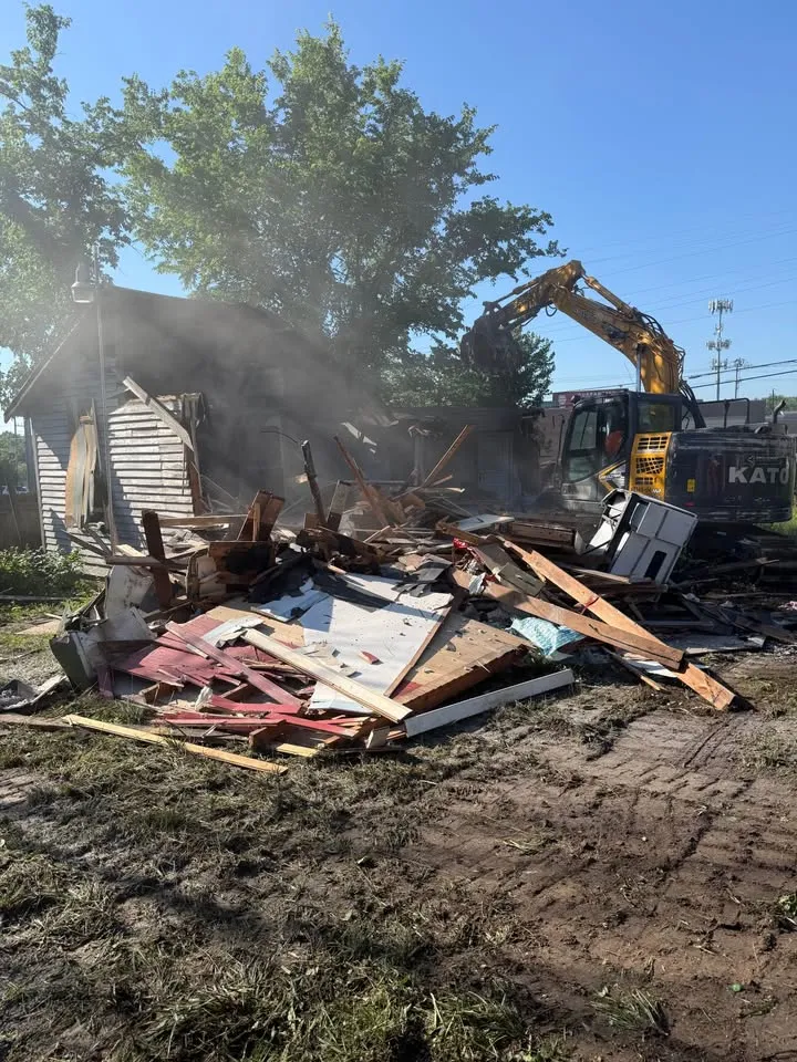 Excavator raising debris over a collapsed timber house, with dust in the air