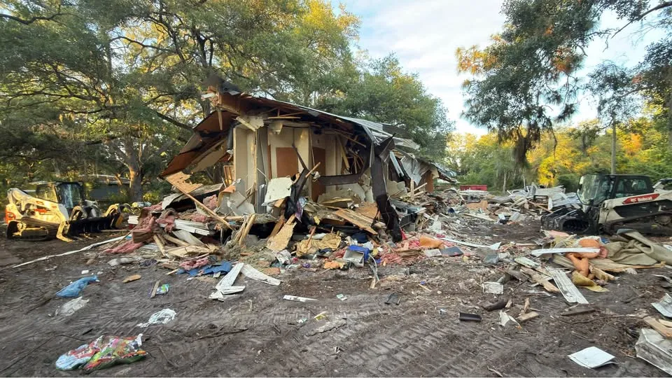 Wide view of a residential teardown with debris spread across the lot and machines at the edges