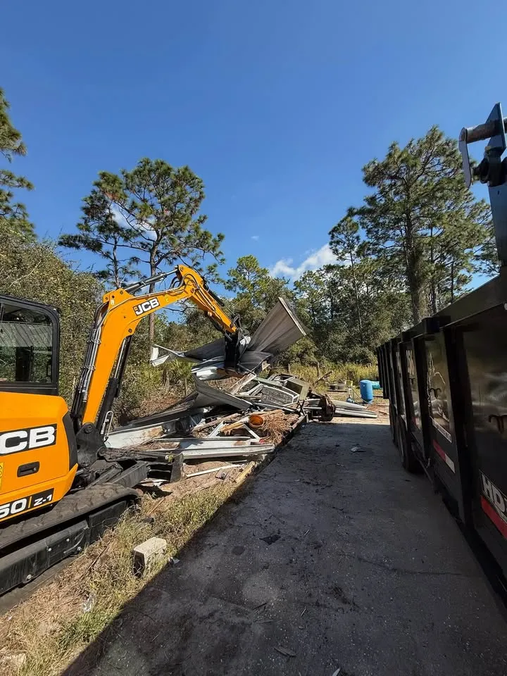 Mini excavator loading house debris into a long dump trailer beside a paved lane