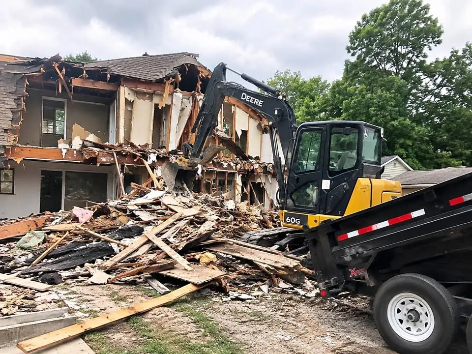 Excavator tearing into a two-storey house while a dump trailer waits beside the pile