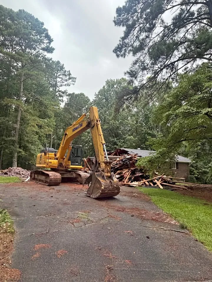 Excavator at the end of a long driveway, taking down a house surrounded by tall trees