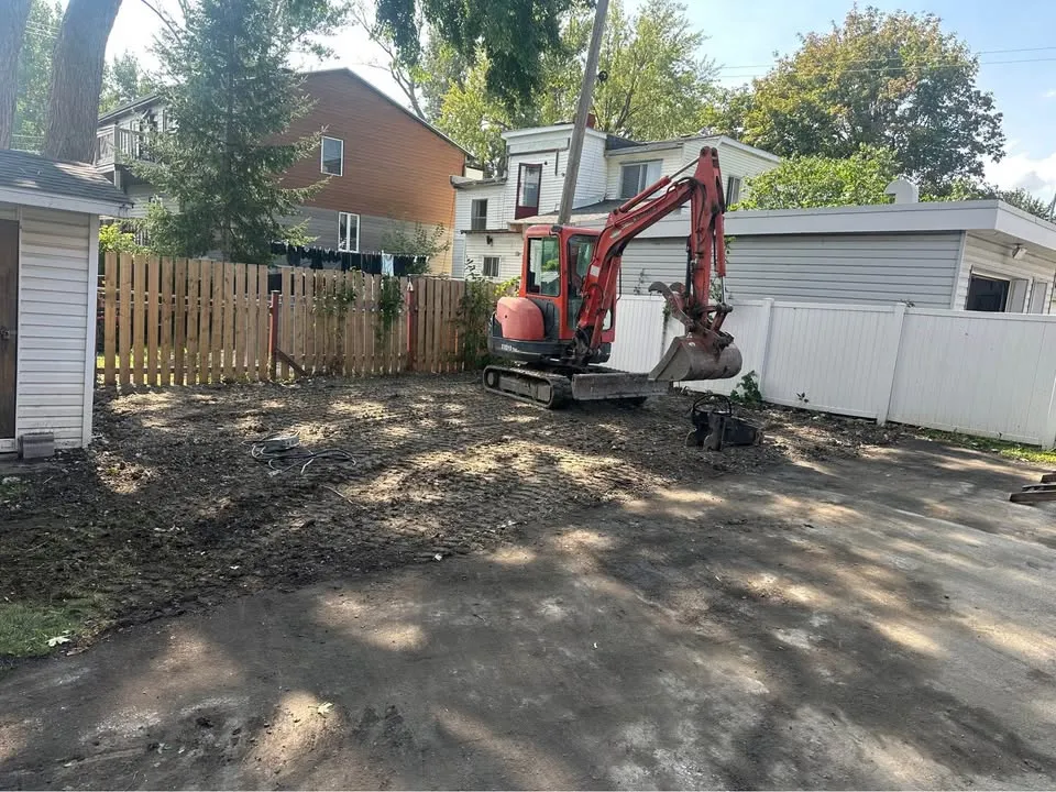 Mini excavator grading a cleared lot between a wood fence and a vinyl fence
