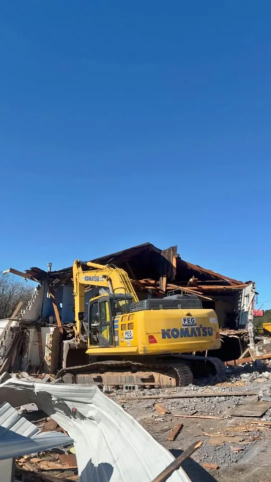 Yellow excavator pulling down the timber roof structure of a large building, with twisted metal cladding in the foreground